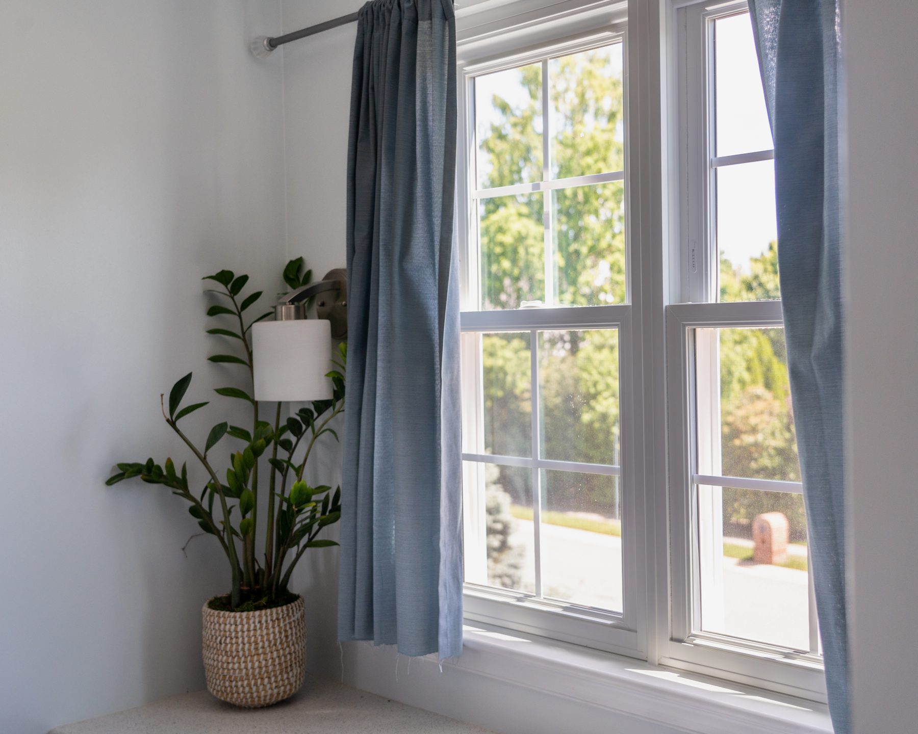 Two white Vytex Fortis double-hung windows with colonial grids and blue linen curtains in a sunlit room
